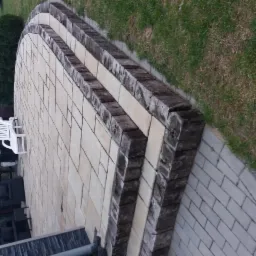 Półokrągły taras z jasnej kostki brukowej z trzema poziomami obrzeży z ciemnego drewna, widok z góry, w tle trawnik i krzewy.