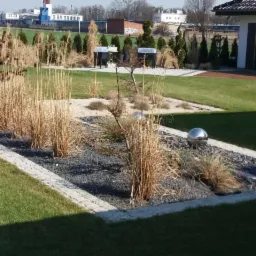 Nowoczesny ogród z rabatą wysypaną czarnym grysem, obrzeżem z kostki i ozdobnymi trawami w odcieniach beżu i brązu, z kulą ze stali nierdzewnej oraz budynkiem w tle.