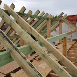 Konstrukcja drewniana dachu w trakcie budowy, widoczne impregnowane belki w kolorze zielonym, płyty OSB i fragment czerwonej dachówki na sąsiednim budynku.