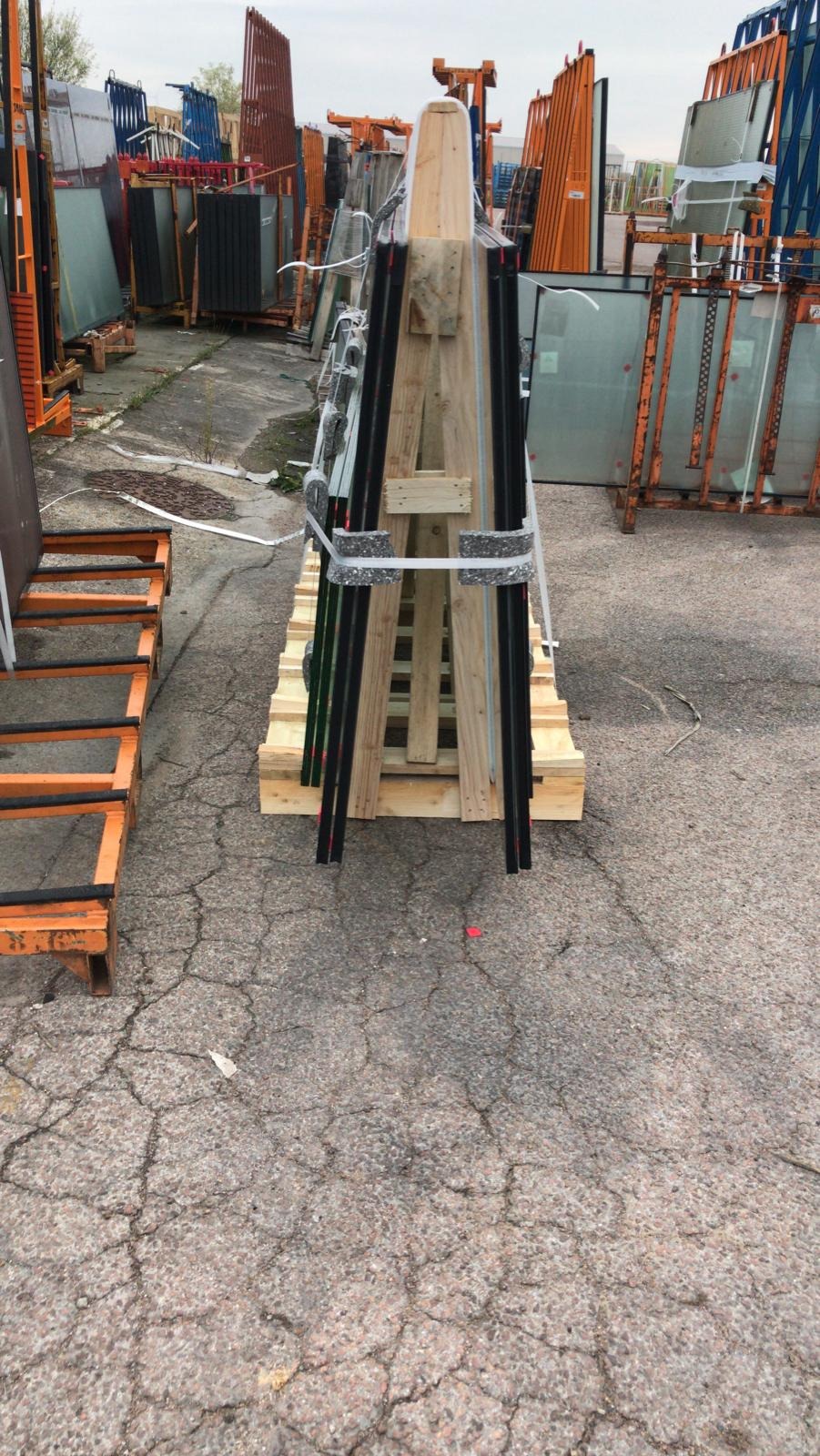 Szyby okienne przygotowane do transportu, zabezpieczone na drewnianej palecie z dodatkowym drewnianym stelażem, widoczne inne materiały budowlane w tle.