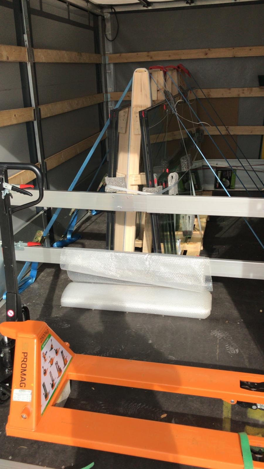 Wnętrze przestrzeni ładunkowej z zabezpieczonymi elementami szklanymi, spiętymi pasami transportowymi, widoczny wózek paletowy PROMAG 2500kg.