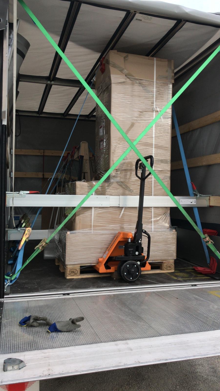 Wnętrze busa transportowego z zabezpieczonym ładunkiem na palecie, z użyciem wózka paletowego i pasów mocujących; widoczne rękawice robocze na podłodze.