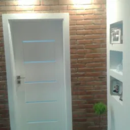 Białe drzwi wewnętrzne z poziomymi, błękitnymi akcentami, osadzone w białej futrynie, na tle ściany z cegły i białej ściany z wnękami na zdjęcia i dekoracje, oświetlone punktowymi lampami sufitowymi.