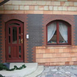 Ciemnoczerwone drzwi wejściowe z przeszkleniami i okrągłe okno z białymi firankami w elewacji z cegły klinkierowej w dwóch odcieniach, przed wejściem mały wiadro i fragment dekoracji z gałązek.