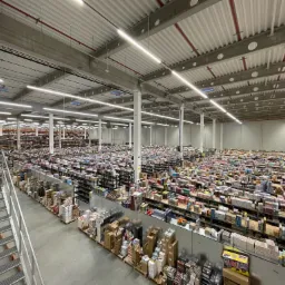 Szeroki plan wnętrza magazynu z wysokim sufitem z widocznymi instalacjami, regałami wypełnionymi towarami i schodami po lewej stronie.