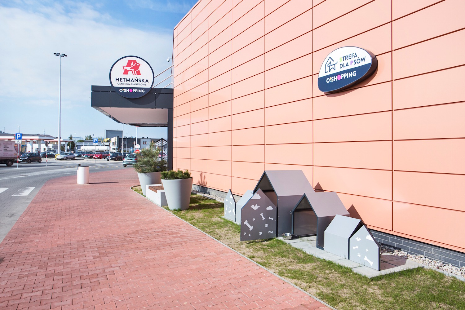 Elewacja budynku usługowego z logo centrum handlowego i strefą dla psów, obejmująca budy w różnych rozmiarach z motywami kości i łap, ustawione na trawniku.
