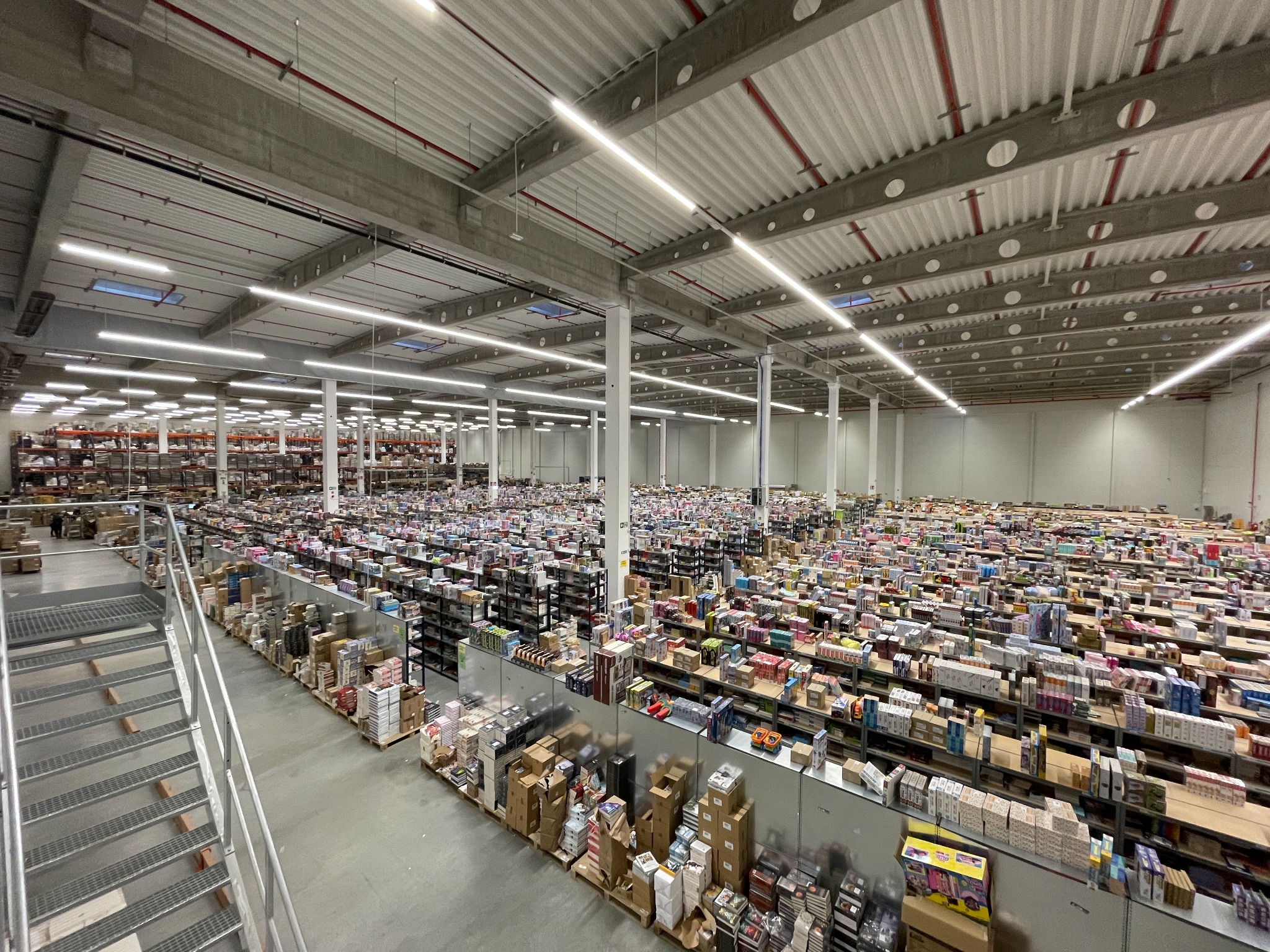 Szeroki plan wnętrza magazynu z wysokim sufitem z widocznymi instalacjami, regałami wypełnionymi towarami i schodami po lewej stronie.