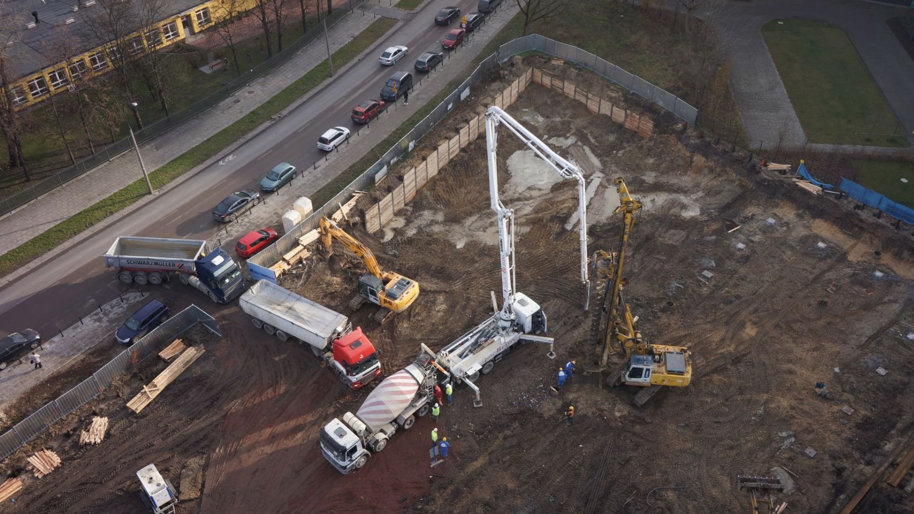 Widok z lotu ptaka na plac budowy z ciężarówkami, betoniarką, pompą do betonu i koparkami, przygotowującymi teren pod fundamenty budynku usługowego w słoneczny dzień.