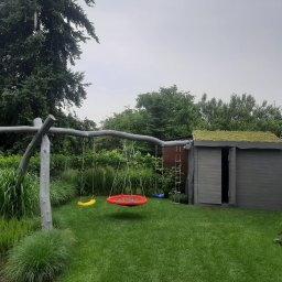 Kater Paweł Marciniak - Ogród z drewnianą huśtawką, kolorowymi siedziskami i szarą drewutnią z zielonym dachem porośniętym roślinnością.