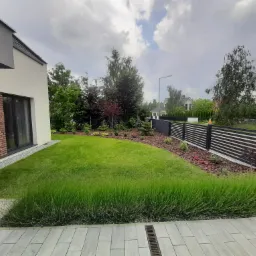 Nowoczesny ogród przydomowy z równo przystrzyżonym trawnikiem, rabatami z korą i niskimi krzewami, obrzeża z szarej kostki brukowej i minimalistyczne ogrodzenie z poziomych desek.