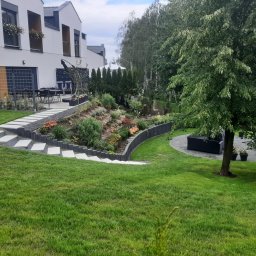 Kater Paweł Marciniak - Nowoczesny ogród przydomowy z tarasem, schodami z płyt betonowych i rabatami z roślinami ozdobnymi, obrzeżami z palisady i strefą relaksu pod drzewem.