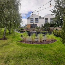 Kater Paweł Marciniak - Nowoczesny ogród przydomowy z trawnikiem, rabatami z ozdobnymi trawami i ścieżką z kamiennymi stopniami prowadzącą do domu o białej elewacji, oświetlony girlandami żarówek.