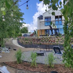 Kater Paweł Marciniak - Prace ziemne w ogrodzie z tarasami obsadzonymi kwiatami, widoczne schody, palenisko na żwirze i pracownik z taczką na tle domu w trakcie budowy.