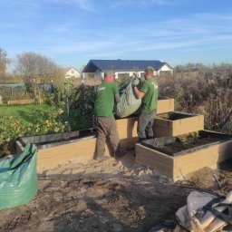 Kater Paweł Marciniak - Dwóch pracowników w zielonych koszulkach z logo firmy przenosi duży worek z ziemią do podwyższonych grządek z imitacji drewna na tle domu jednorodzinnego.