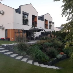 Kater Paweł Marciniak - Nowoczesny ogród przydomowy z betonowymi schodami prowadzącymi na trawnik i taras z parasolem, rośliny ozdobne na zboczu, białe domy z kwiatami w oknach.