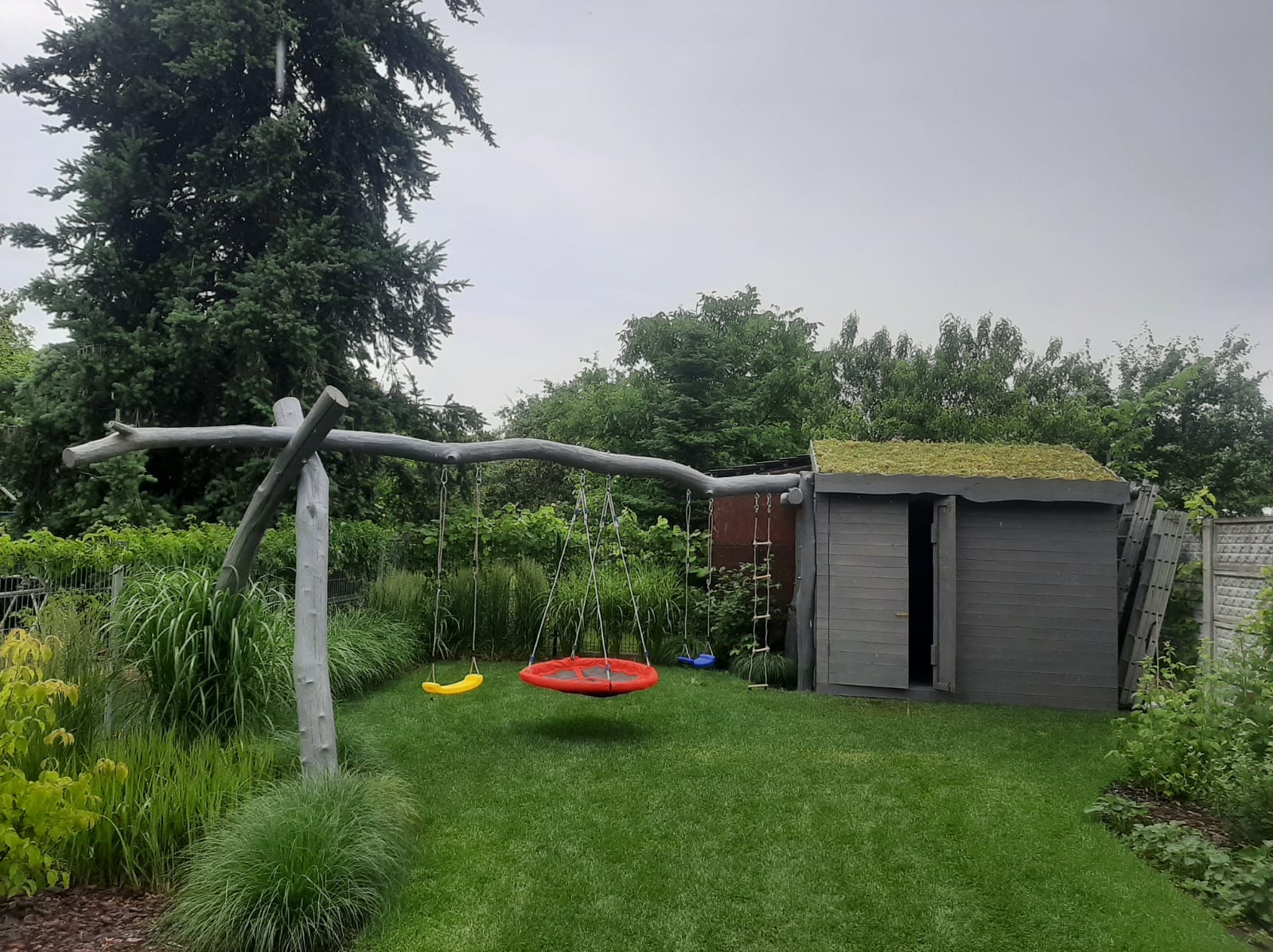 Ogród z drewnianą huśtawką, kolorowymi siedziskami i szarą drewutnią z zielonym dachem porośniętym roślinnością.