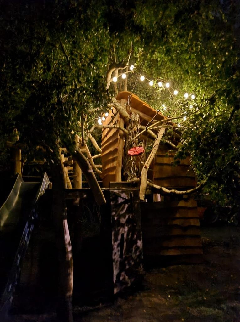 Oświetlony domek na drzewie z huśtawką i zjeżdżalnią, otoczony zielenią, w nocnej scenerii.