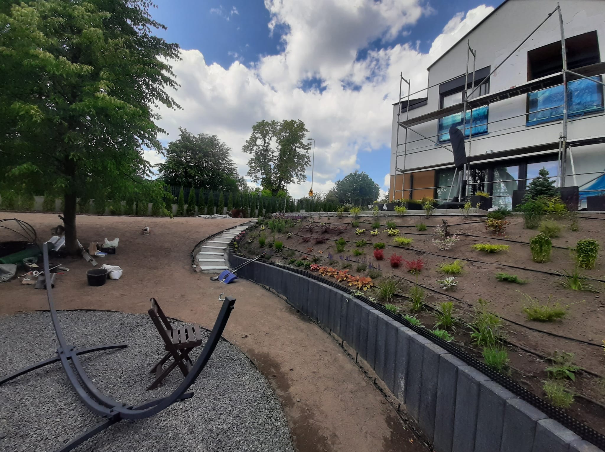 Nowo założony ogród z podwyższonymi rabatami, ścieżką i schodami, widoczne elementy systemu nawadniania, w tle dom w trakcie remontu z rusztowaniami.