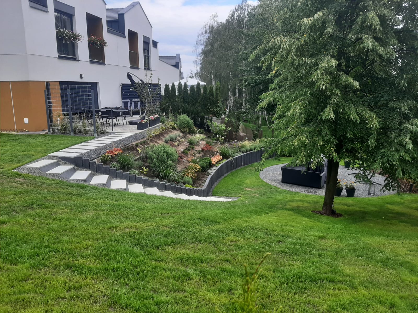 Nowoczesny ogród przydomowy z tarasem, schodami z płyt betonowych i rabatami z roślinami ozdobnymi, obrzeżami z palisady i strefą relaksu pod drzewem.