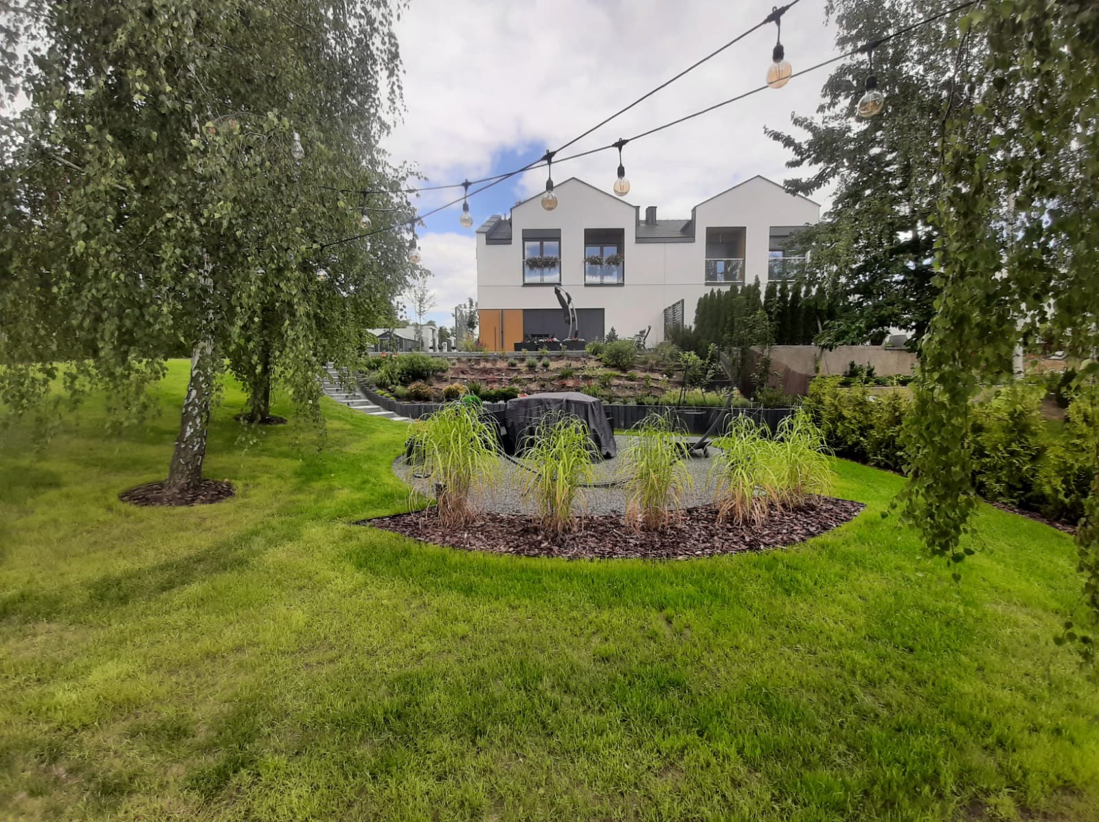 Nowoczesny ogród przydomowy z trawnikiem, rabatami z ozdobnymi trawami i ścieżką z kamiennymi stopniami prowadzącą do domu o białej elewacji, oświetlony girlandami żarówek.