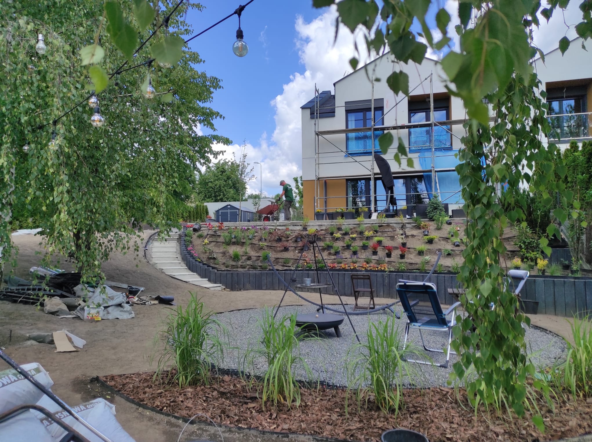 Prace ziemne w ogrodzie z tarasami obsadzonymi kwiatami, widoczne schody, palenisko na żwirze i pracownik z taczką na tle domu w trakcie budowy.