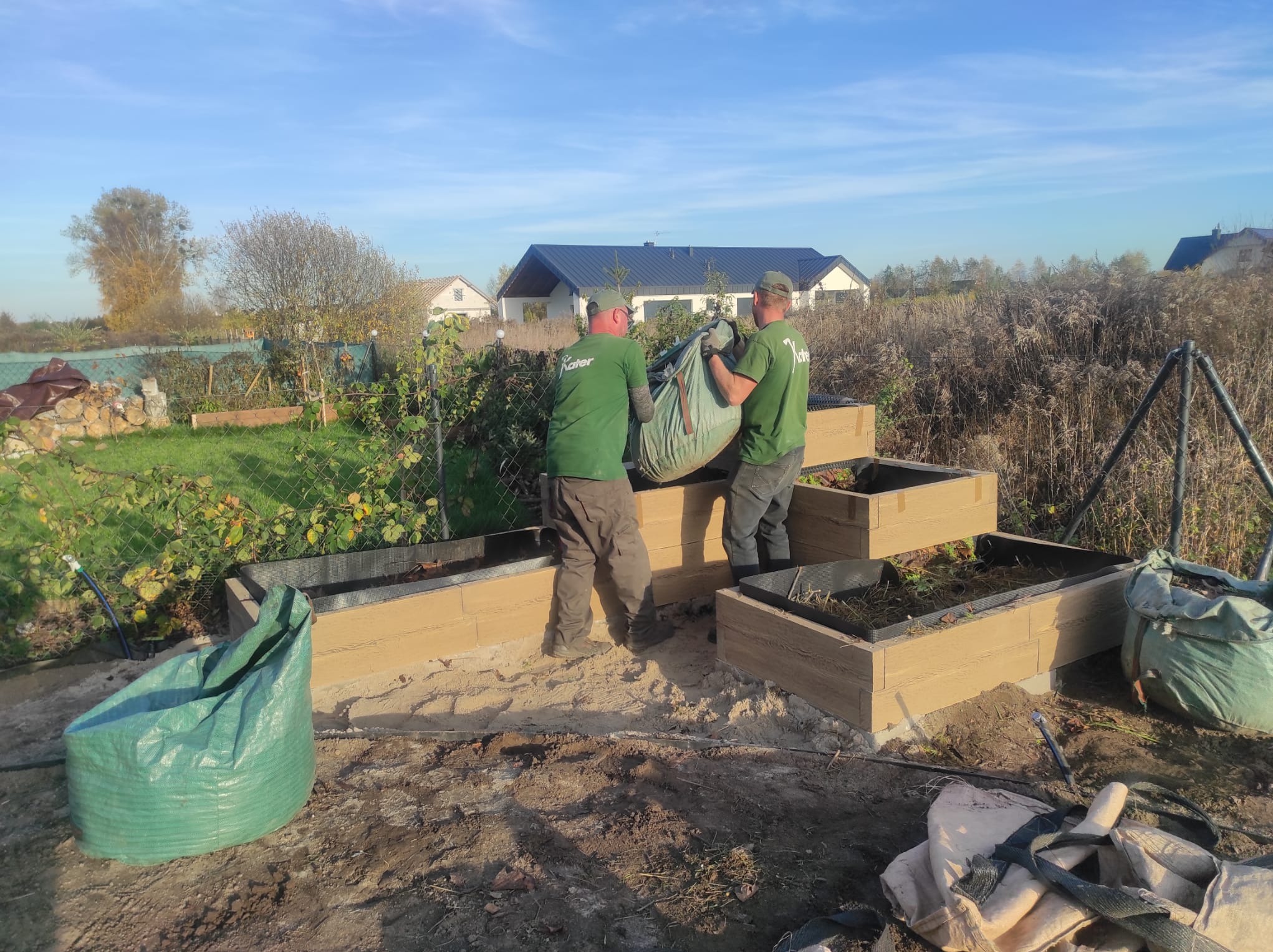 Dwóch pracowników w zielonych koszulkach z logo firmy przenosi duży worek z ziemią do podwyższonych grządek z imitacji drewna na tle domu jednorodzinnego.