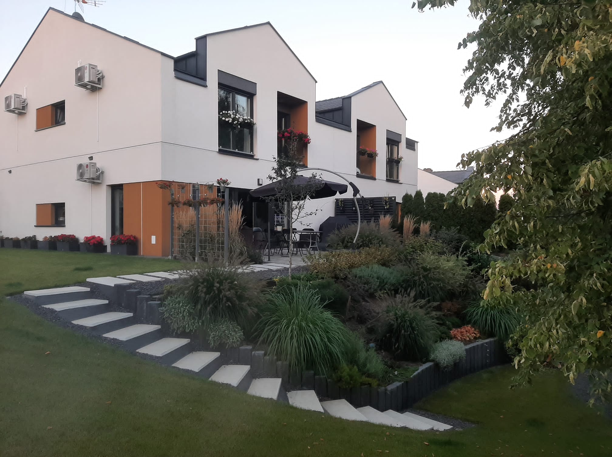 Nowoczesny ogród przydomowy z betonowymi schodami prowadzącymi na trawnik i taras z parasolem, rośliny ozdobne na zboczu, białe domy z kwiatami w oknach.