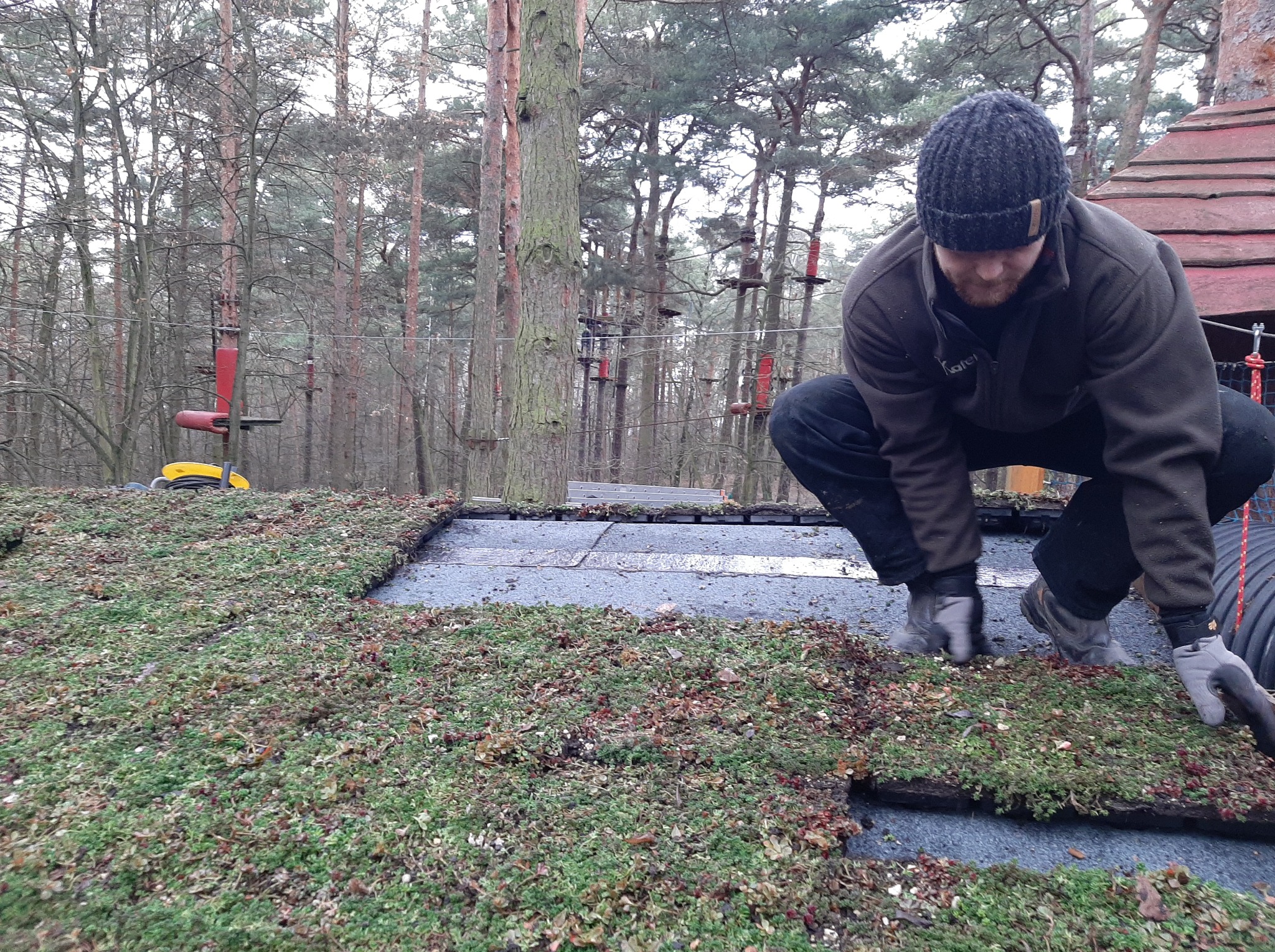 Pracownik w czapce i rękawicach układa segmenty zielonego dachu na płaskiej powierzchni, w tle las i elementy parku linowego.
