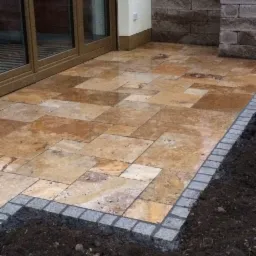 Ułożony taras z trawertynu z obrzeżem z kostki granitowej, widok na przesuwne drzwi tarasowe i fragment ściany z dekoracyjnej cegły.