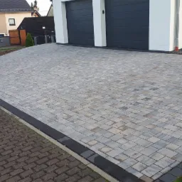 Podjazd do garażu wyłożony szarą kostką o nieregularnym wzorze, obrzeża wykończone czarnym elementem, widok z góry.