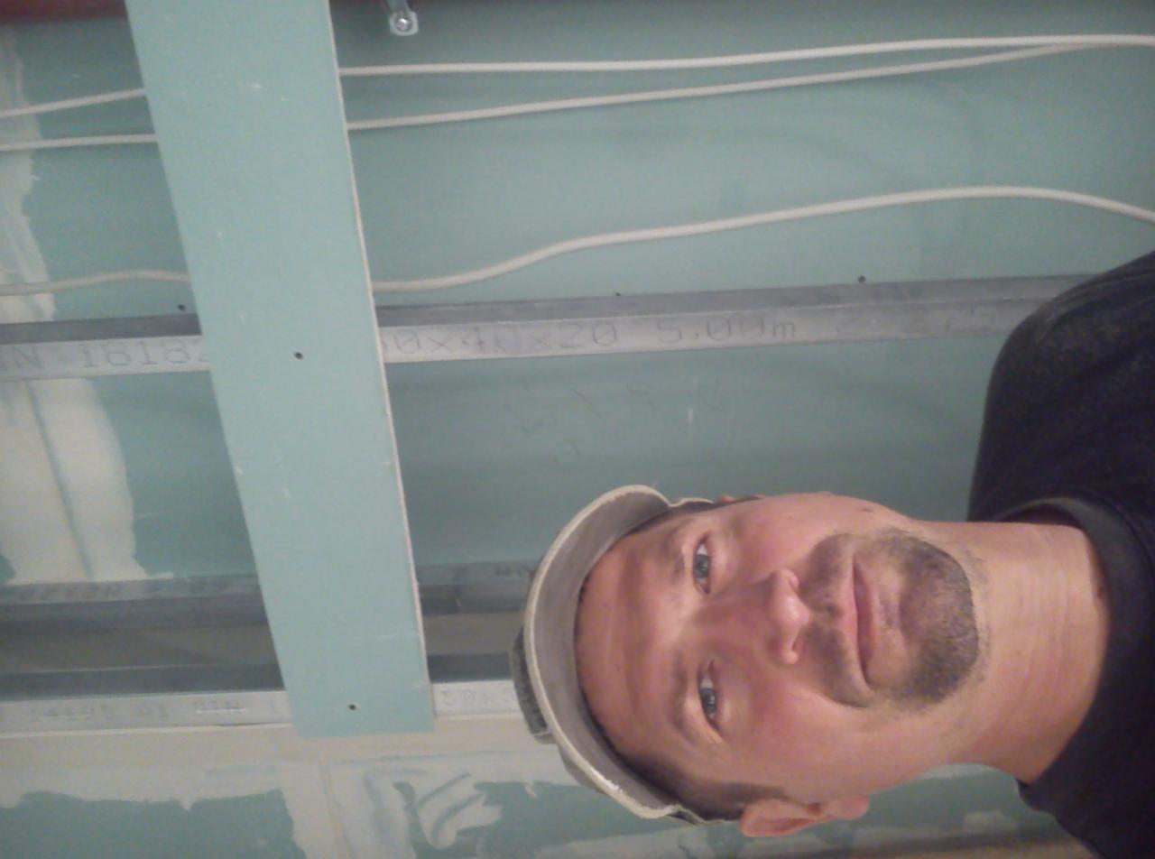 Mężczyzna w kasku spogląda w dół, na tle konstrukcji sufitu z płyt kartonowo-gipsowych, widoczne profile metalowe i fragmenty płyt.