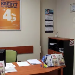 Biuro kredytowe z plakatem informującym o nisko oprocentowanym kredycie gotówkowym od 4,9%, kalendarzem i certyfikatami na ścianie, biurkiem z komputerem, broszurami i doniczką z zieloną rośliną.