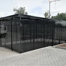 Nowoczesna, ażurowa konstrukcja z czarnego metalu, prawdopodobnie wiata śmietnikowa lub rowerowa, na tle betonowej kostki brukowej i placu budowy z żurawiem.