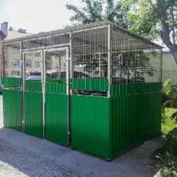 Zadaszona wiata śmietnikowa z zielonymi panelami i metalową konstrukcją, widoczna na tle bloku mieszkalnego i drzew.
