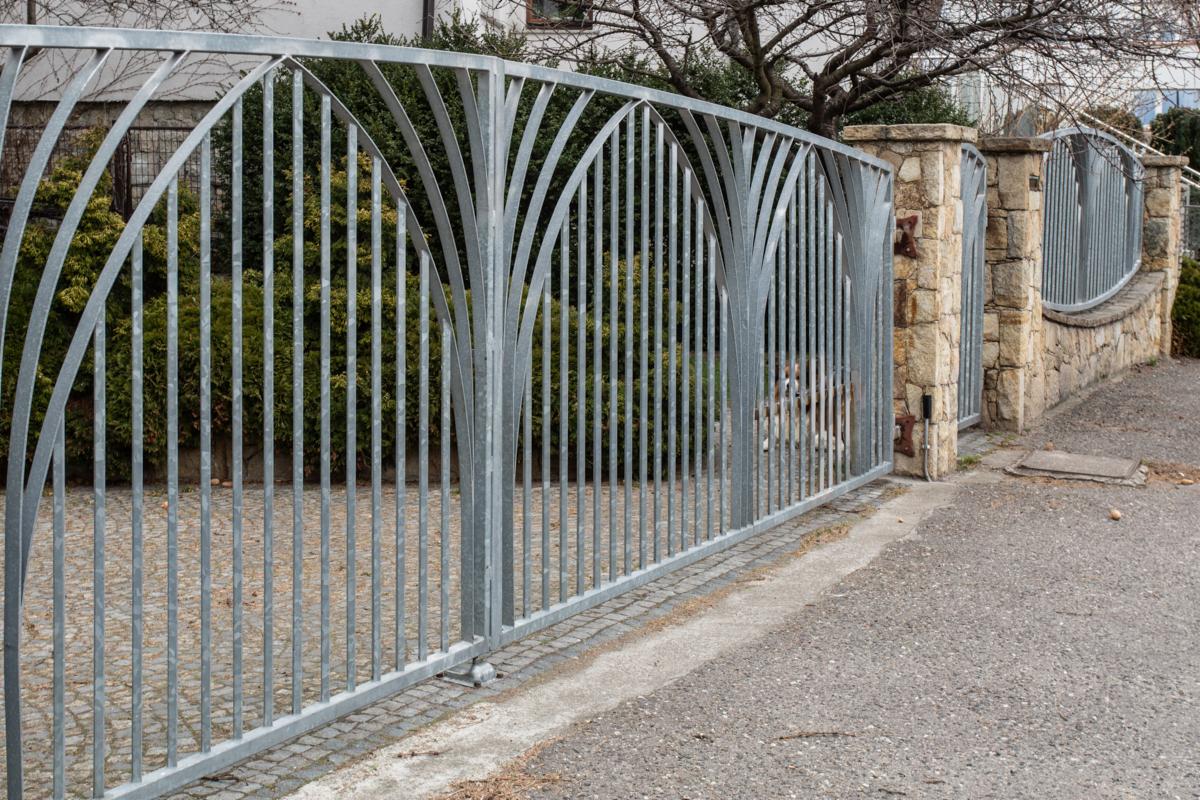 Metalowa brama wjazdowa z łukowatym wzorem, osadzona między kamiennymi słupkami, widok na posesję z krzewami i drzewami.