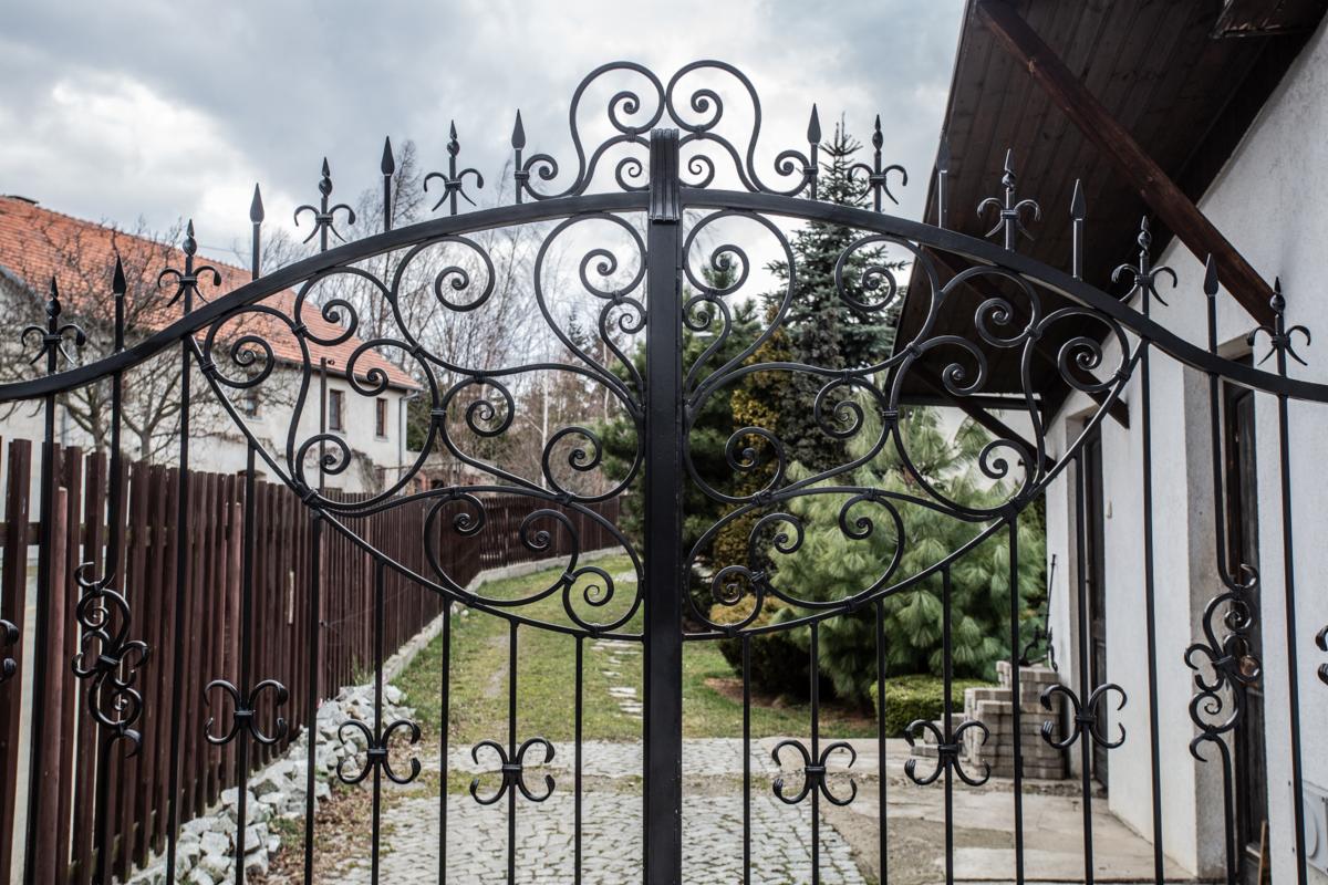 Kuta brama wjazdowa z ozdobnymi elementami w kształcie spirali i kwiatów, widok na posesję z brukowaną ścieżką i domem w tle.