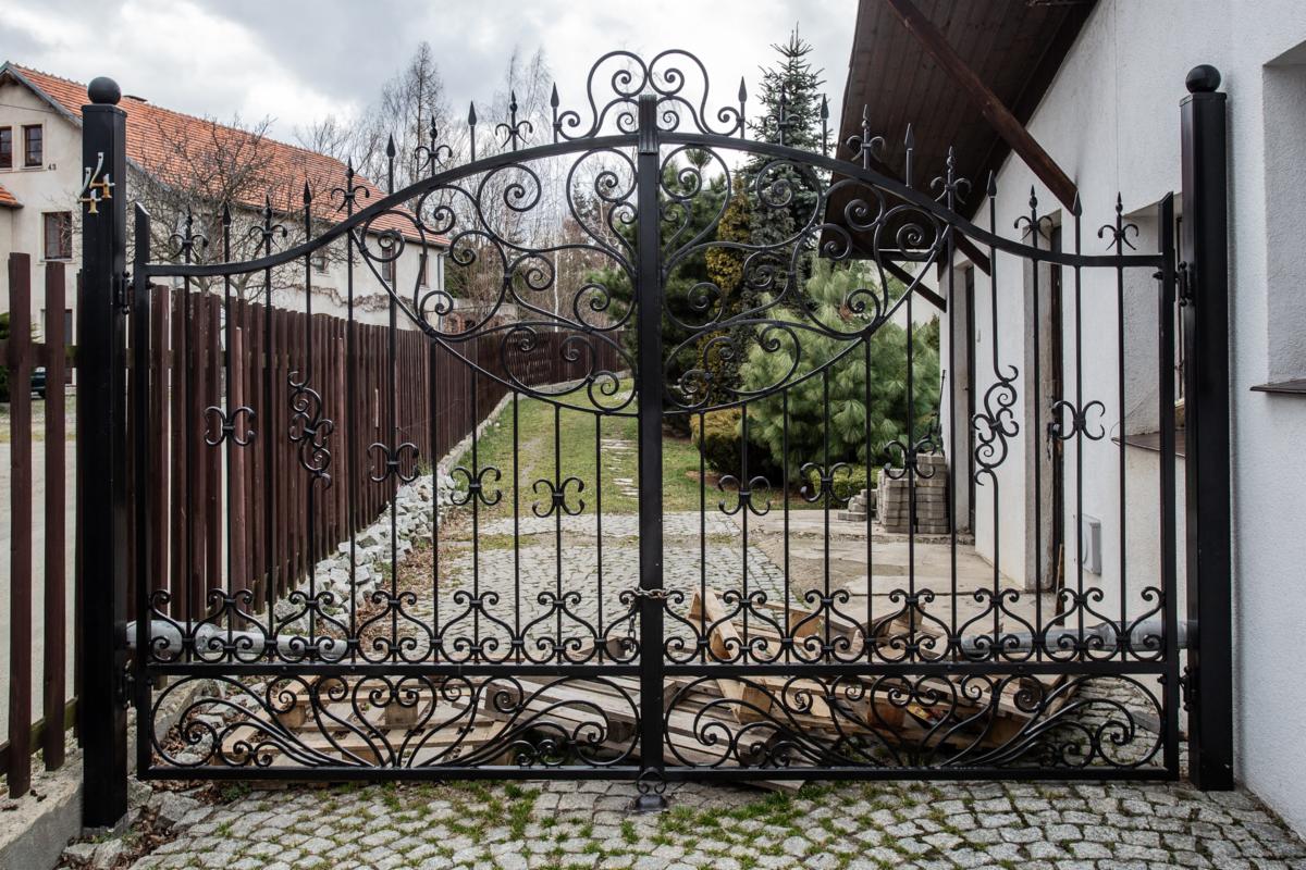Kuta, czarna brama wjazdowa z ozdobnymi elementami na tle brukowanej ścieżki i zieleni ogrodu, zamknięta na łańcuch z kłódką.