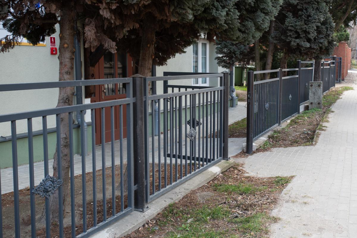 Długi, szary płot z metalowych prętów z ozdobnymi rozetami, biegnący wzdłuż chodnika i budynku z brązowymi drzwiami, w tle drzewa iglaste.