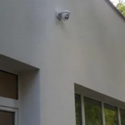 Biała kamera monitoringu kopułkowa zamontowana na elewacji budynku z widocznymi oknami, ujęcie z dołu.