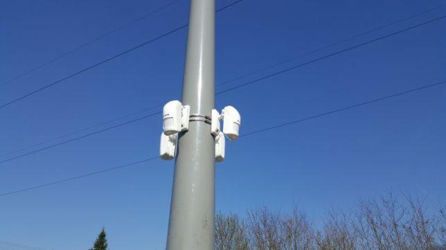 Szary słup oświetleniowy z zamontowanymi czterema białymi czujnikami ruchu, zabezpieczonymi metalową obejmą, na tle błękitnego nieba i linii energetycznych.