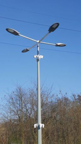 Wysoki, szary słup oświetleniowy z czterema ramionami zakończonymi nowoczesnymi lampami LED, dodatkowo wyposażony w kamery monitoringu, na tle błękitnego nieba i drzew bez liści.