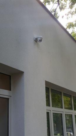 Biała kamera monitoringu kopułkowa zamontowana na elewacji budynku z widocznymi oknami, ujęcie z dołu.