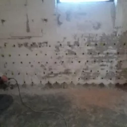 Pracownik wierci otwory w wilgotnej, ceglanej ścianie, przygotowując ją do osuszania metodą iniekcji krystalicznej. Widoczne nawiercone otwory i ślady wilgoci na ścianie.
