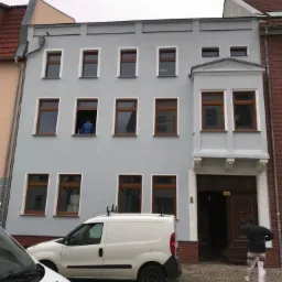 Odnowiona, trzypiętrowa kamienica z szarą elewacją i brązowymi oknami, białe obramowania okien, biały van firmowy zaparkowany przed budynkiem, pracownik w oknie na drugim piętrze, drugi wchodzi...