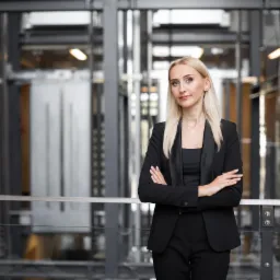 Profesjonalna blondynka w czarnym garniturze, skrzyżowane ręce, nowoczesne biuro w tle z widoczną windą panoramiczną.