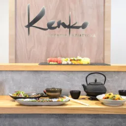 Aranżacja stołu w restauracji azjatyckiej: sushi, sałatki, czarki na sake i czajnik do herbaty, w tle logo restauracji 'Kenko Kuchnia Azjatycka'.