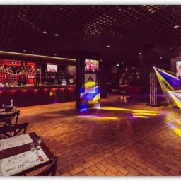 Wnętrze lokalu z barem, stolikami z menu i oświetleniem dyskotekowym, widoczny ekran z tańczącymi osobami.