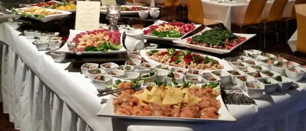 Bogato zastawiony stół cateringowy z różnorodnymi przekąskami, w tym sałatkami, przystawkami w małych miseczkach i talerzem z łososiem i nachos, całość na białym obrusie z dekoracyjnymi fałdami.