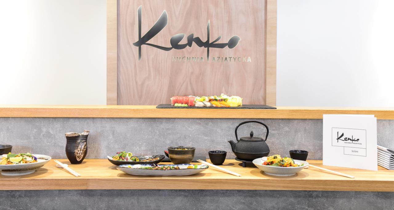 Aranżacja stołu w restauracji azjatyckiej: sushi, sałatki, czarki na sake i czajnik do herbaty, w tle logo restauracji 'Kenko Kuchnia Azjatycka'.