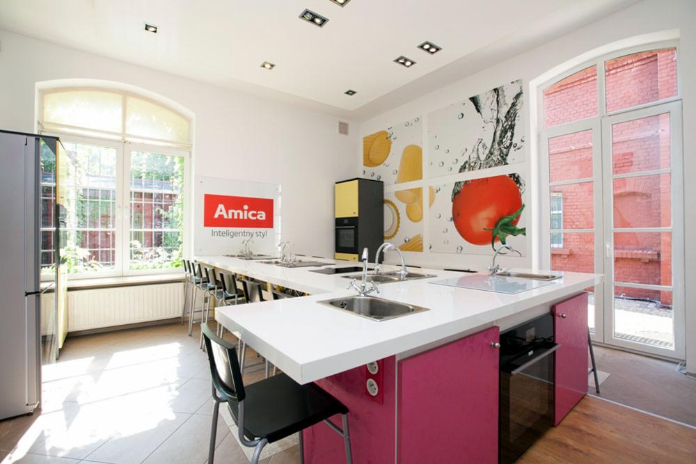 Duża, jasna kuchnia z długim blatem i kilkoma zlewozmywakami, plakatem z motywami jedzenia na ścianie i logo Amica, prawdopodobnie kuchnia pokazowa lub sala szkoleniowa.